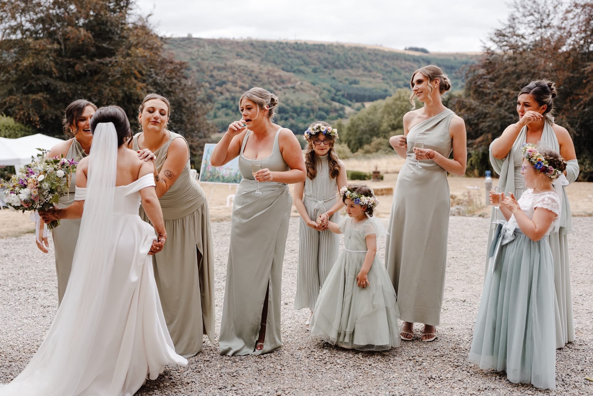Bridesmaids in sage green dresses holding wildflower bouquets in front of English countryside hills