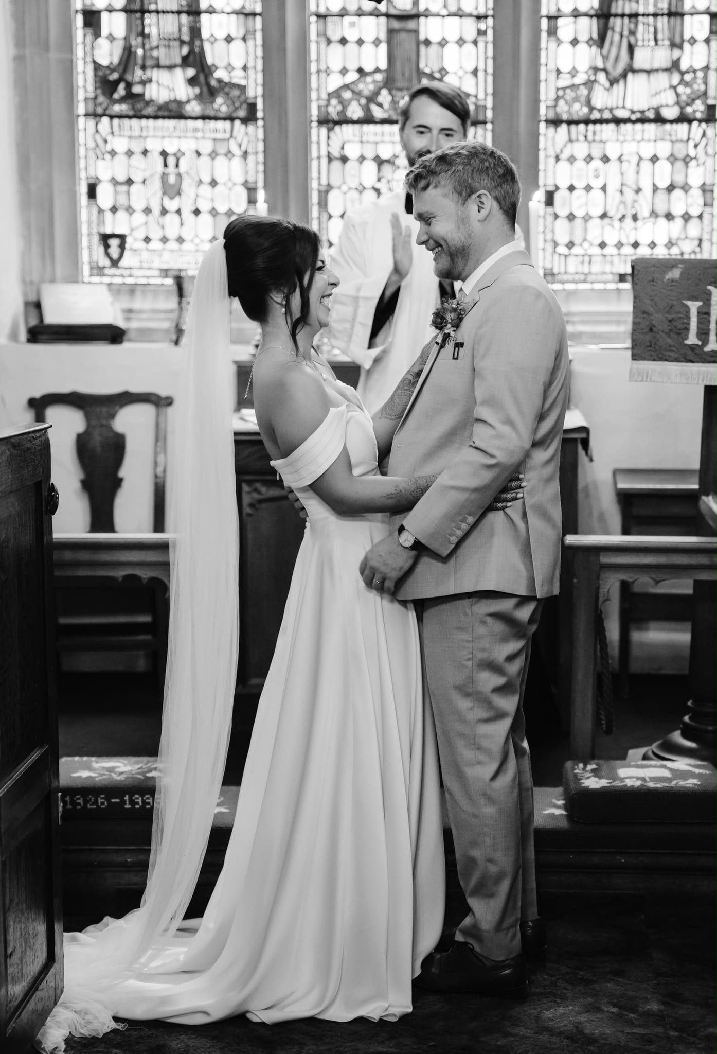 Bride and groom exchanging vows in front of stained glass in a small English stone chapel, black and white