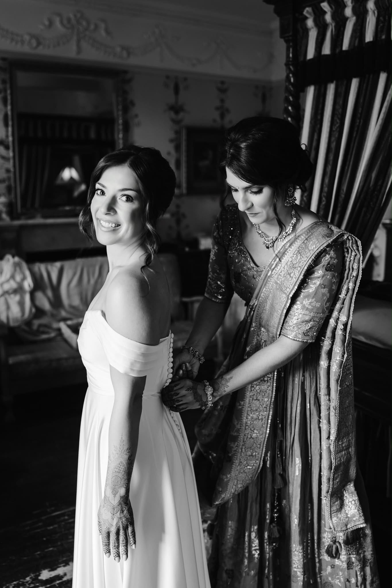 A family member in a gold and rose sari helps the bride into her LALEDA wedding dress