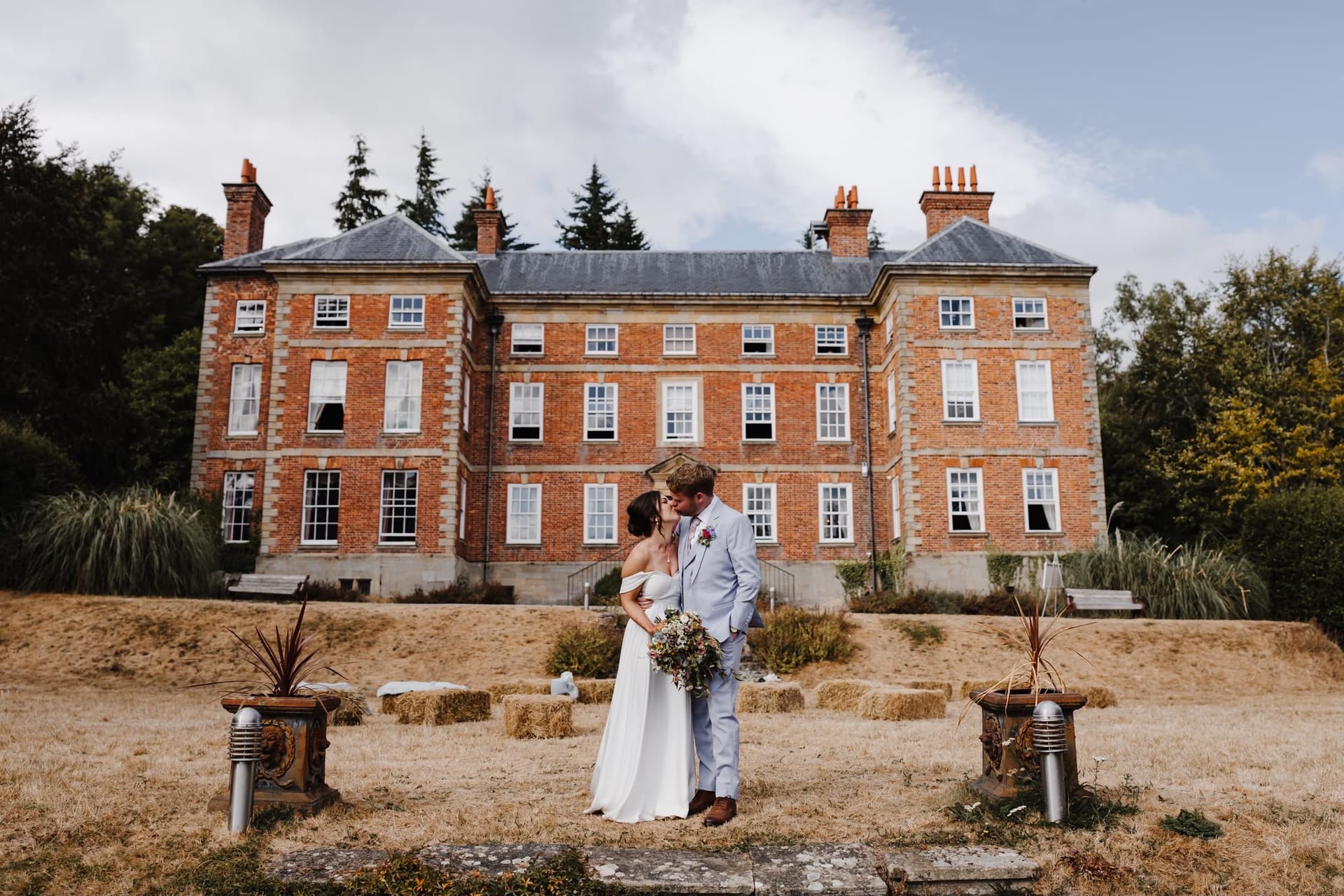 Bride in a plain white LALEDA wedding gown and groom kissing in front of a red-brick English manor house